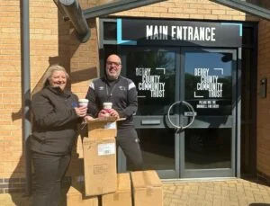 Two people - one female one male - are pictured smiling outside a building with the words Derby County Community Trust on the front.