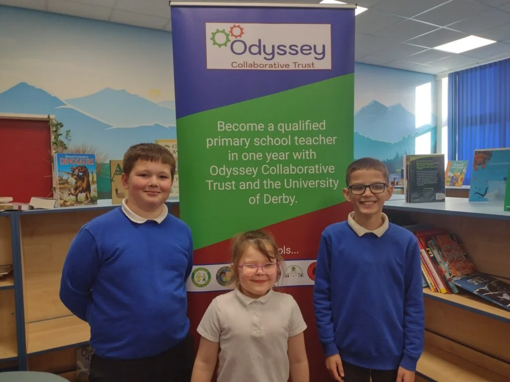 Children stand in front of sign advertising the teacher training course being delivered through Odyssey Collaborative Trust