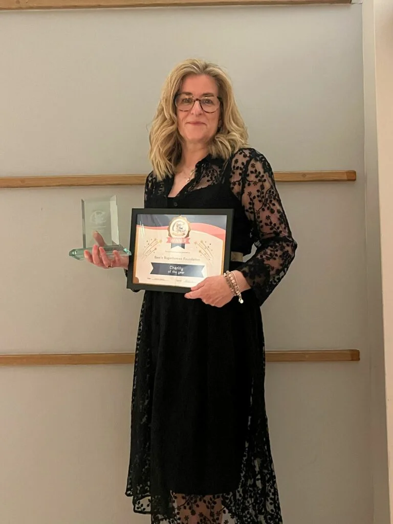 Rachel Liew the founder of Sam's Superheroes Foundation standing in front of a wall holding her award