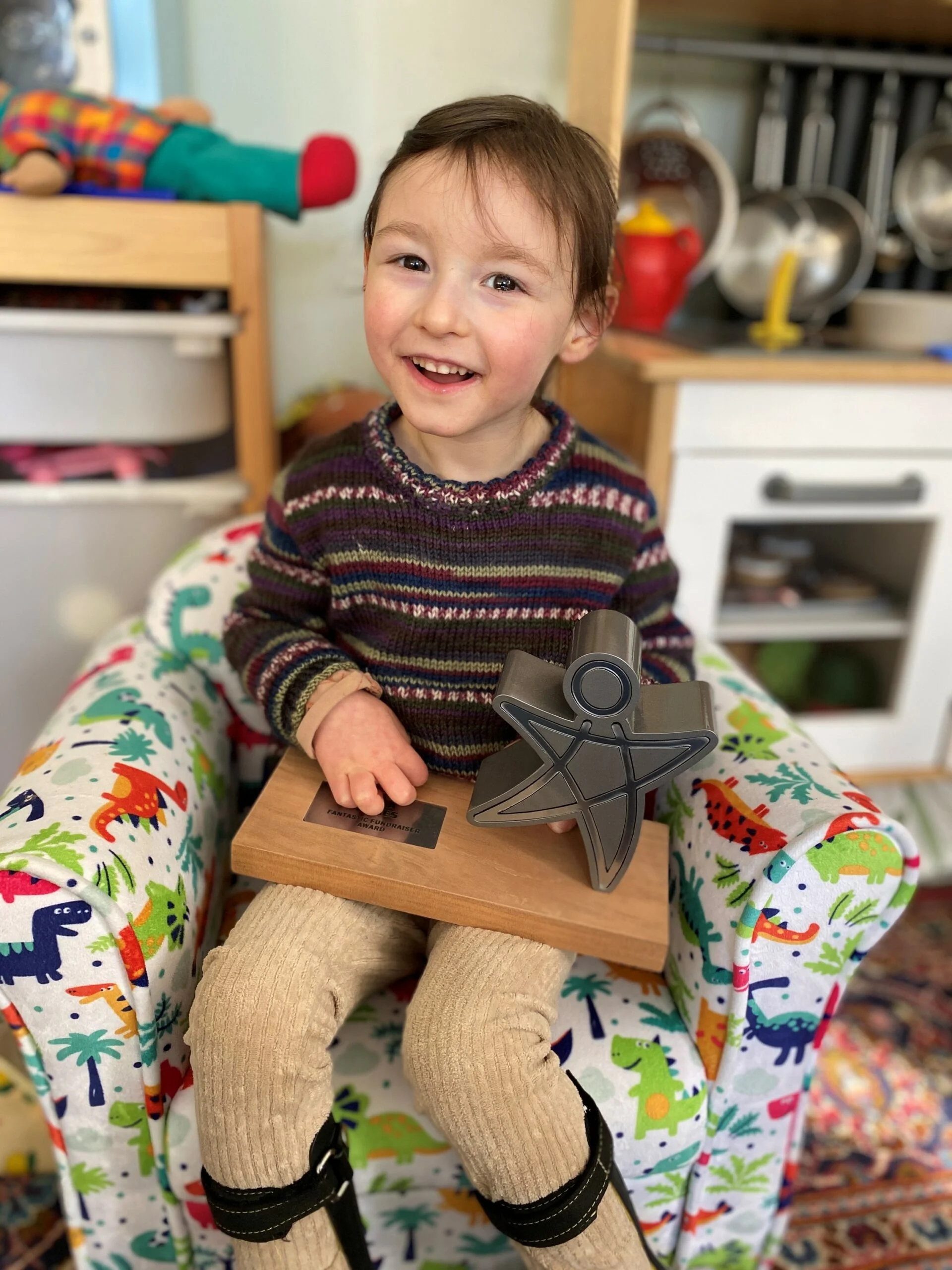 a photo of Lolo who has cerebral palsy sat with her child of courage award on a dinosaur print chair