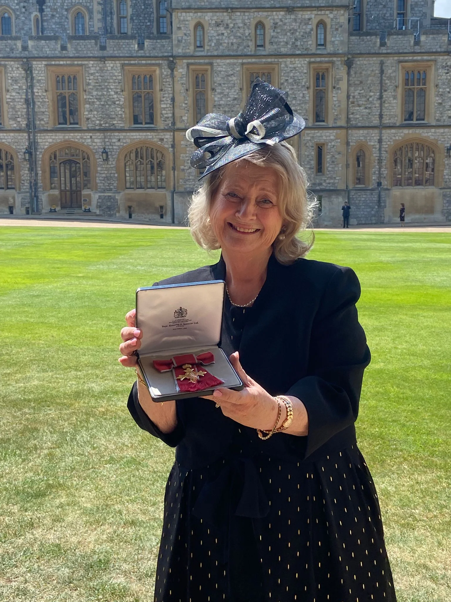 Wendy Daunt holds OBE medal in presentation box in front of palace.